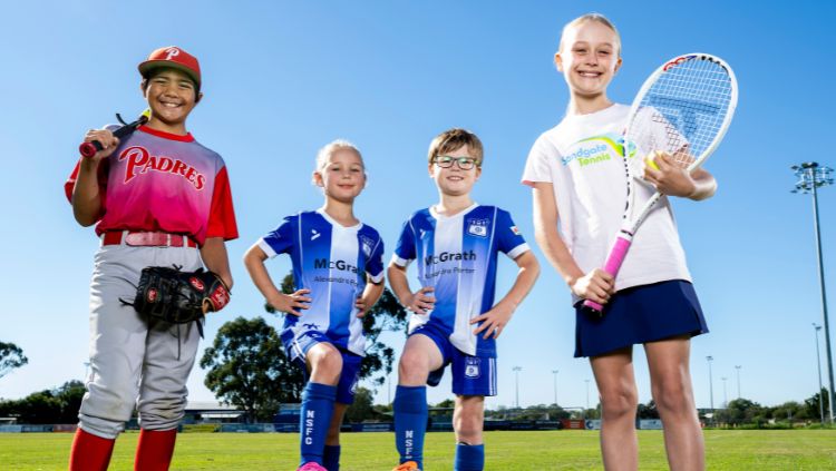 Four children in sports attire standing on a field: one baseball player, two soccer players, and one tennis player.