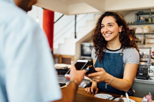 A woman stands behind a counter holding out a card reader while another person holds their phone to the card reader