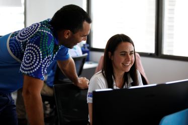 A woman is sitting at a desk looking at a computer screen while a man is leaning over looking at her. The man is wearing a Baidam shirt with Indigenous artwork on the sleeve.