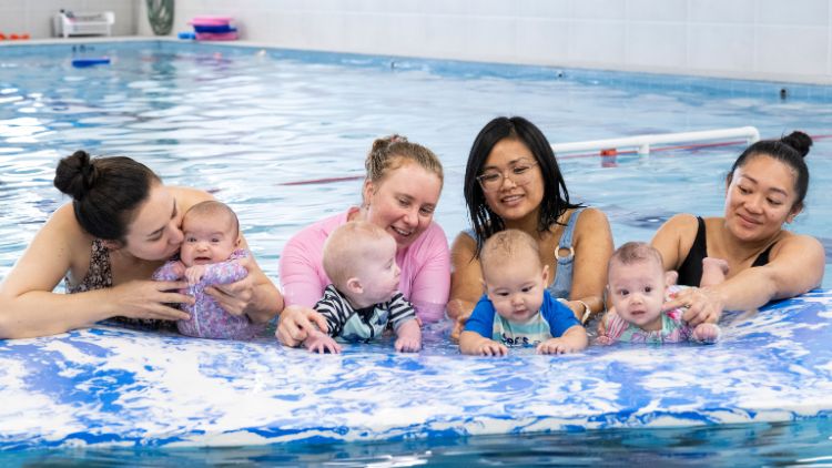 babies swimming atop of floating mat in a pool, being held by adults