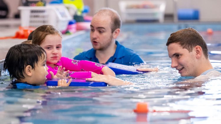 Children with kickboards being taught in the pool how to swim by adults