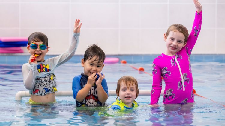 Four children standing in an indoor pool wearing protective swimming suits and goggles.