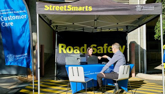 Two people sitting across from each other over a table what has a print and laptop on top of a blue table cloth, under a Street Smarts tent with the back flap down and Road Safety in green on it.