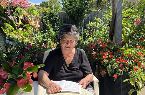old lady sitting at an outdoor table with her garden behind her