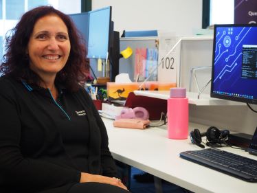 Monique Cooper is a smiling woman with dark hair, wearing a Queensland Government shirt and cardigan and is sitting at a desk with a computer